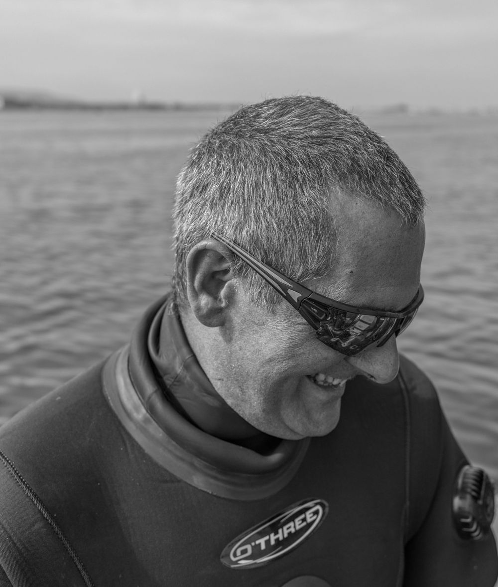 A wetsuit-clad individual smiles while wearing sunglasses near the water's edge in a black and white photograph.