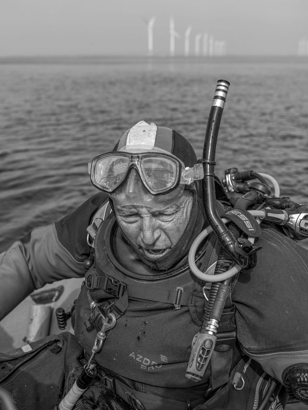 A scuba diver wearing diving gear and goggles in black and white against a misty ocean background.