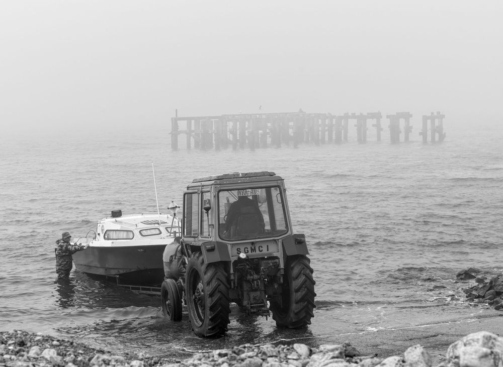 A vintage tractor sits next to a boat on a misty beach with a distant pier visible through the fog.