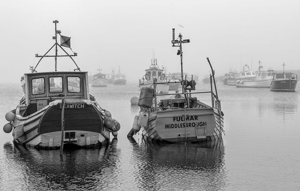 Two small fishing boats moored in foggy harbor with vessels visible in background.