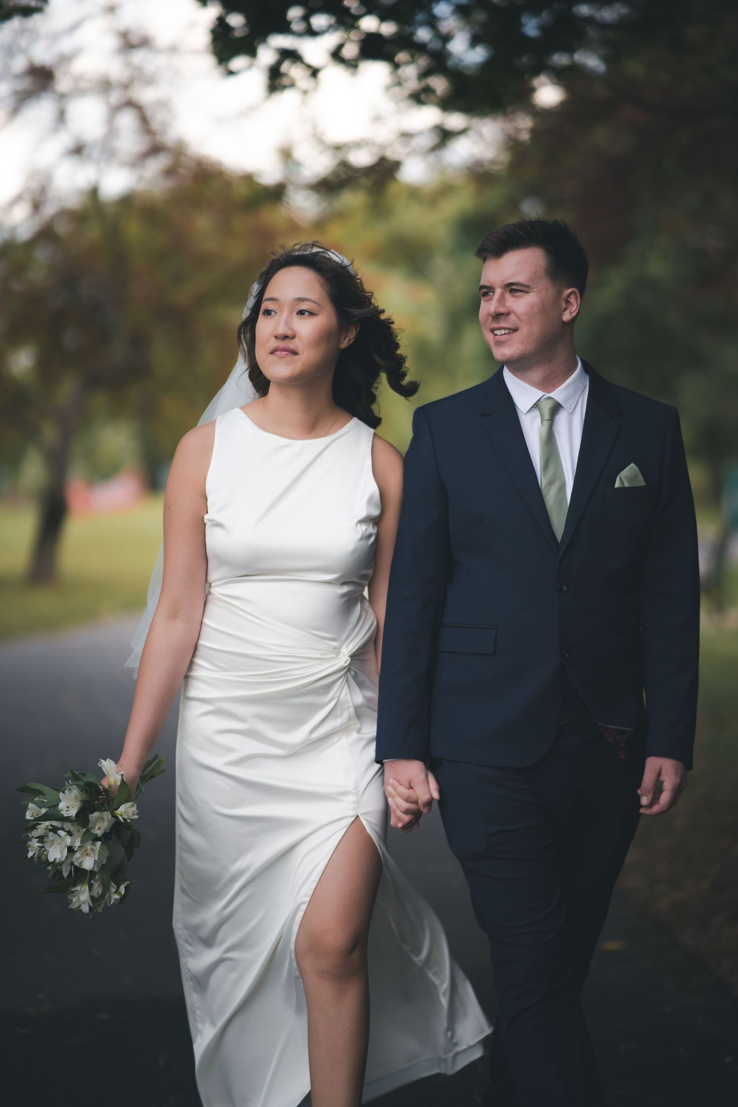 A newlywed couple walks outdoors, with the bride in a white dress with slit and groom in dark suit holding hands.