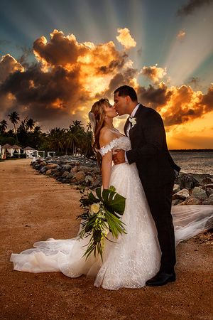 A romantic wedding couple embrace on a tropical beach at sunset with dramatic orange clouds and palm trees in the background. - Puerto Rico