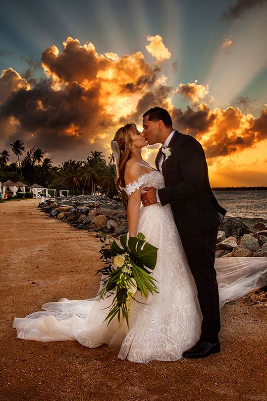 A romantic wedding couple embrace on a tropical beach at sunset with dramatic orange clouds and palm trees in the background. - Puerto Rico