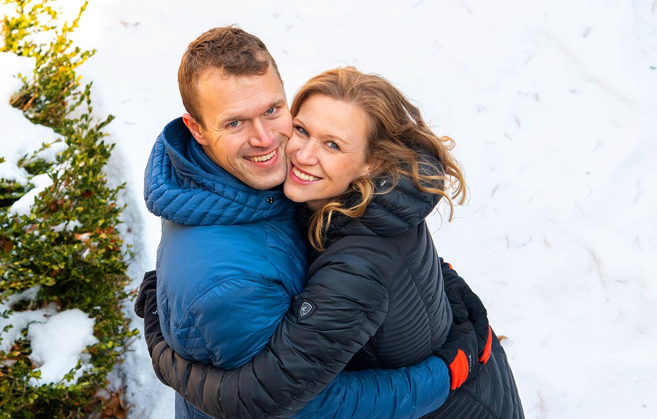 A couple in winter coats share an affectionate embrace outdoors with snow and greenery in the background. (Highland Park, Rochester NY)