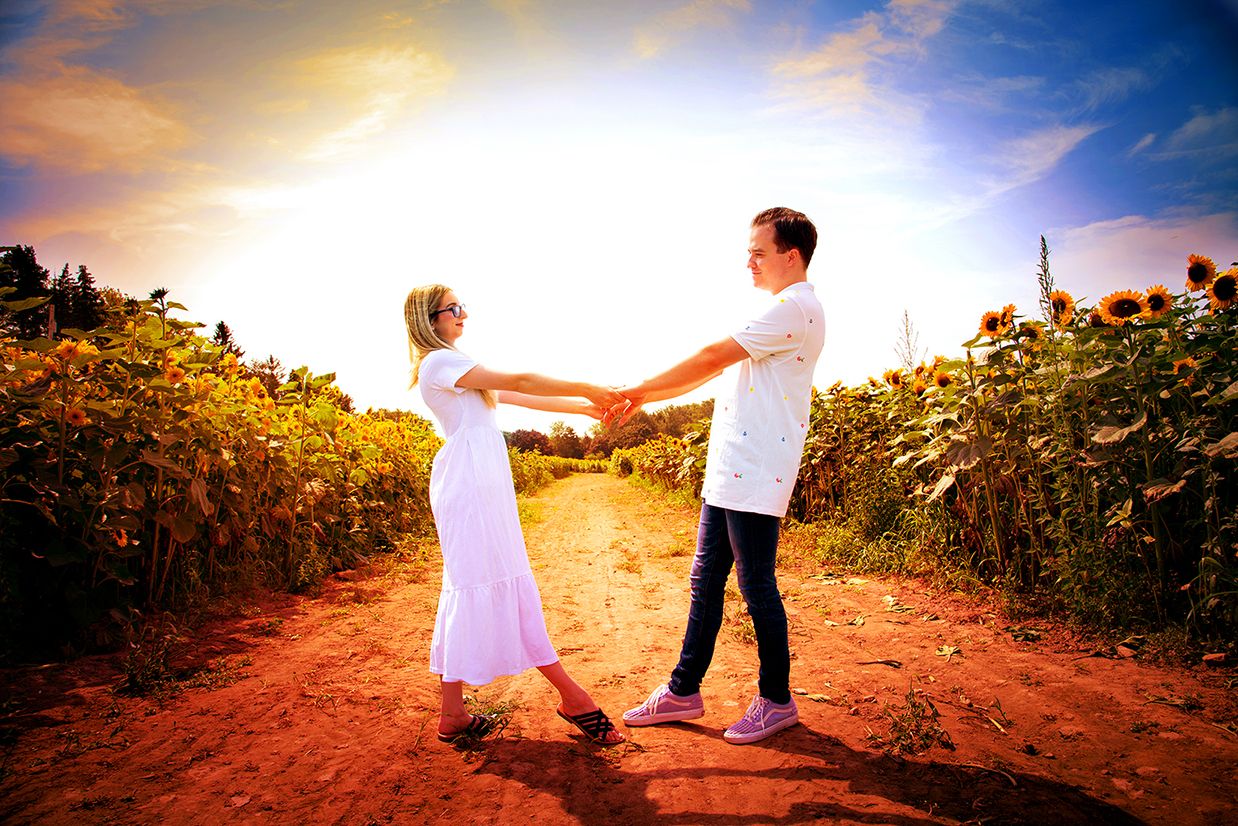 Two people in white outfits dance together on a dirt path at sunset with sunflower fields on both sides. (Wickham Farms Penfield NY)