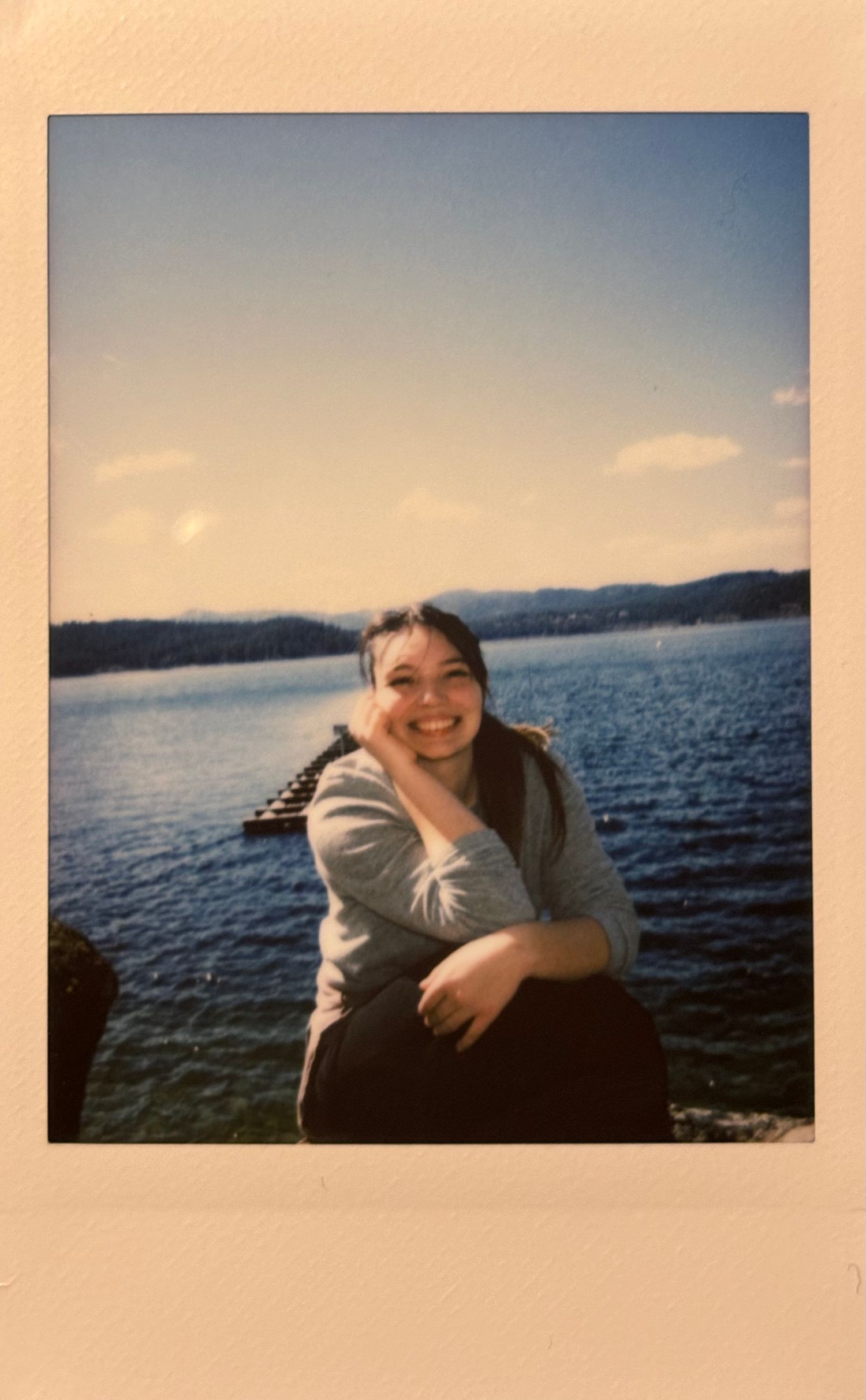 Smiling young woman posing for a Polaroid photo by a scenic mountain lake on a sunny day.