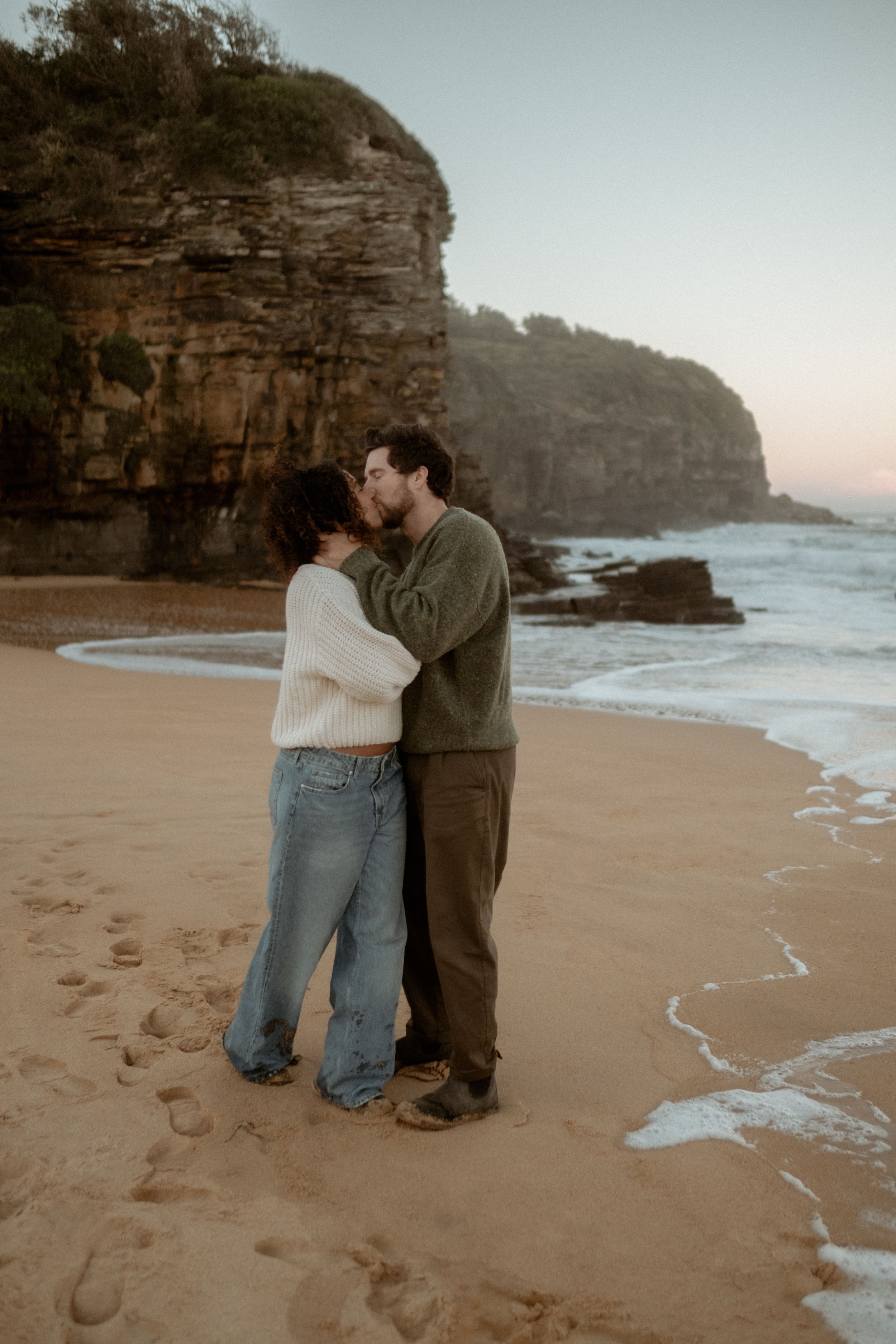 A couple kissing on a sandy beach with cliffs and the ocean behind them.