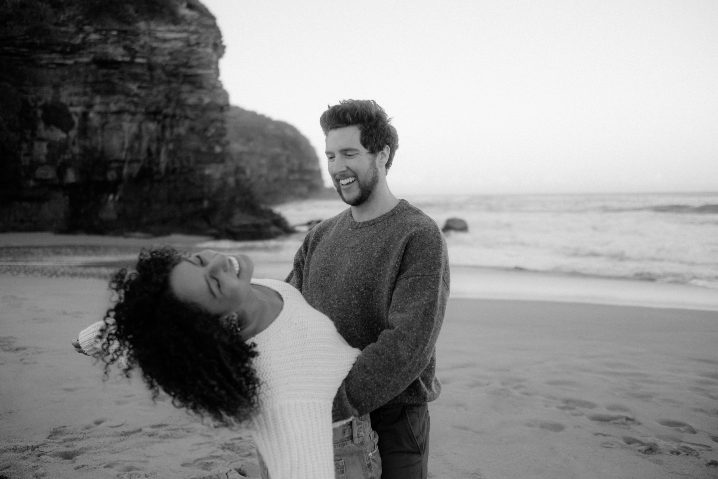 Laughing couple twirling on a sandy beach with the ocean and cliffs behind them