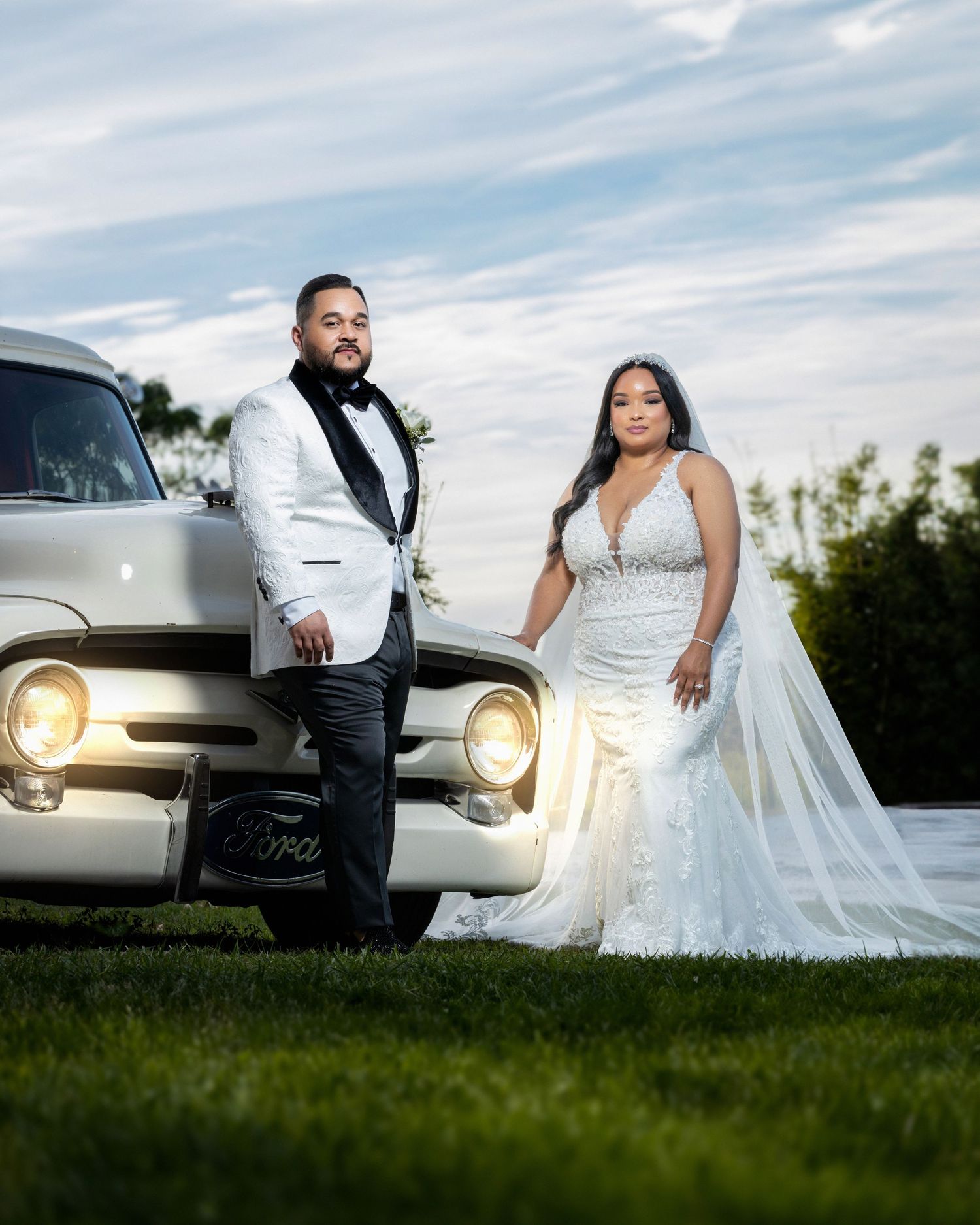 Wedding couple poses with vintage Ford car, groom in white tuxedo and bride in fitted gown.