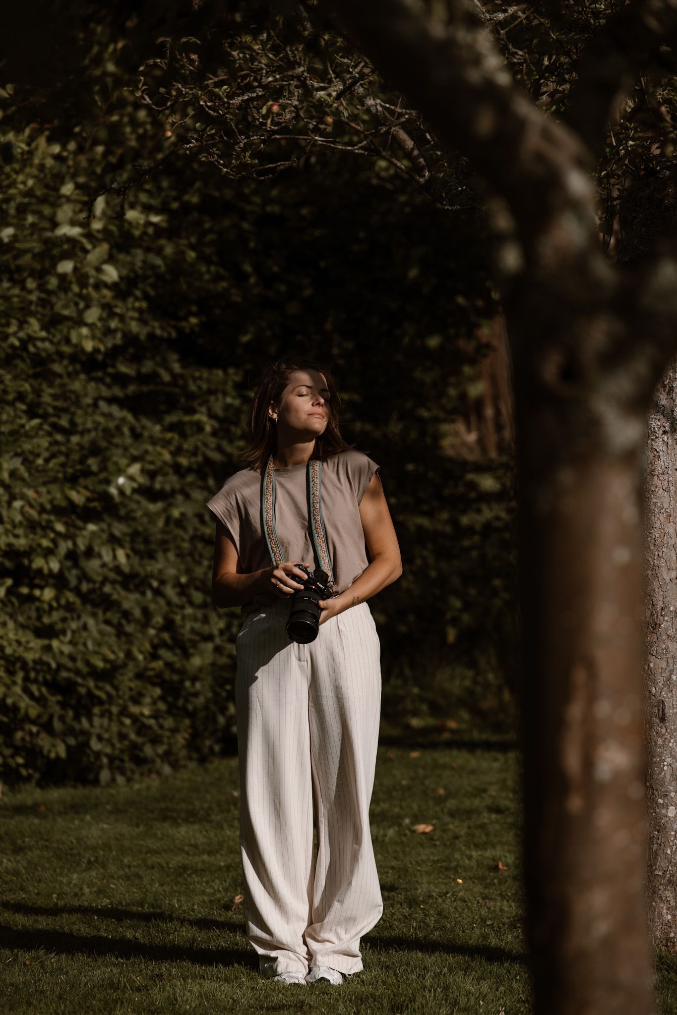 A photographer in a grey t-shirt and white flowy pants stands in dappled sunlight among trees in a garden setting.