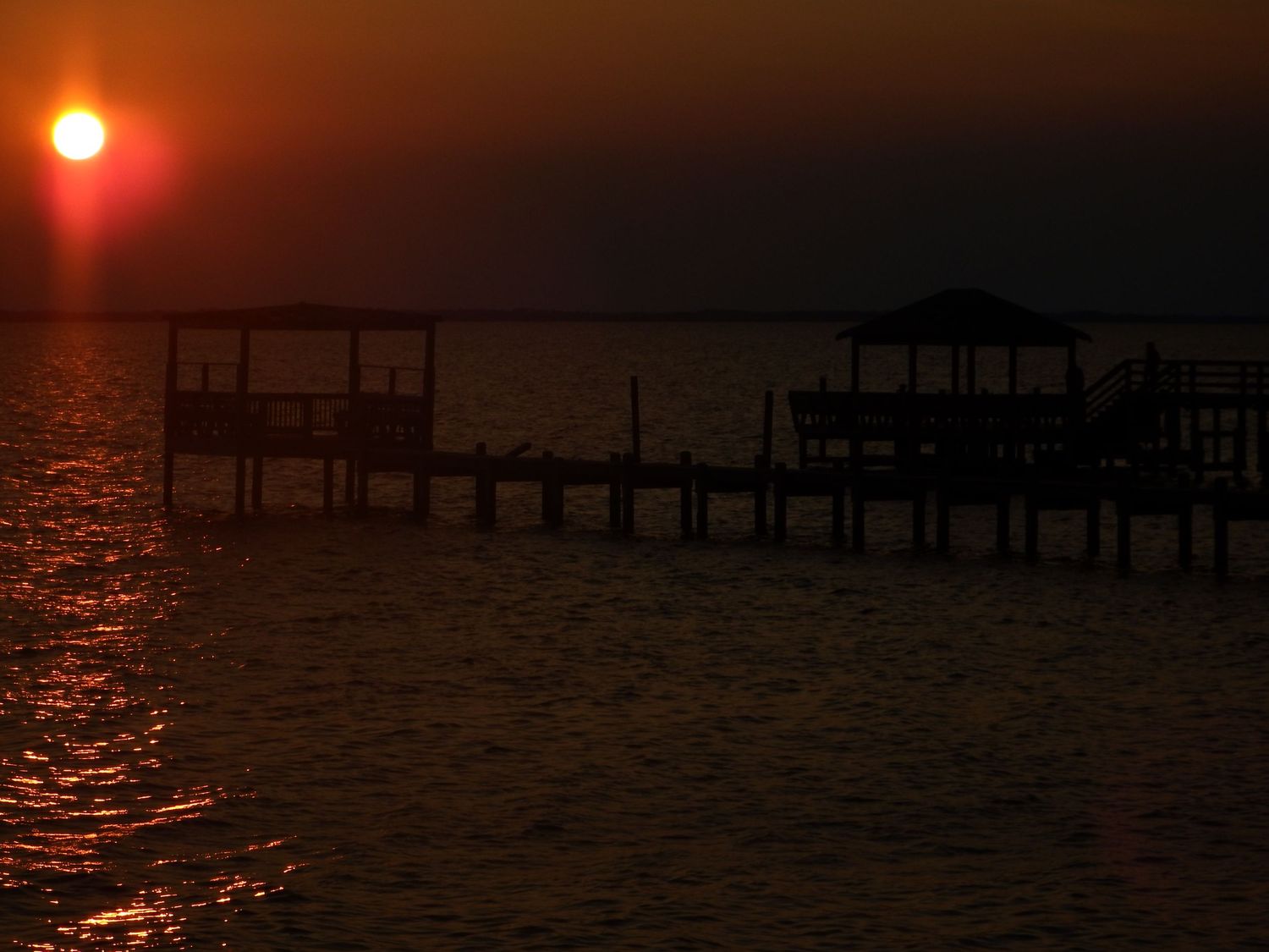 A wooden pier silhouetted against a dramatic red sunset over calm waters.