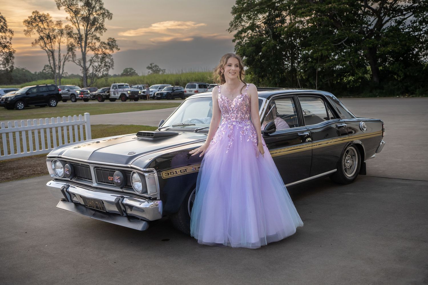 A purple and pink formal gown next to a classic black Ford Falcon at sunset in a parking area.