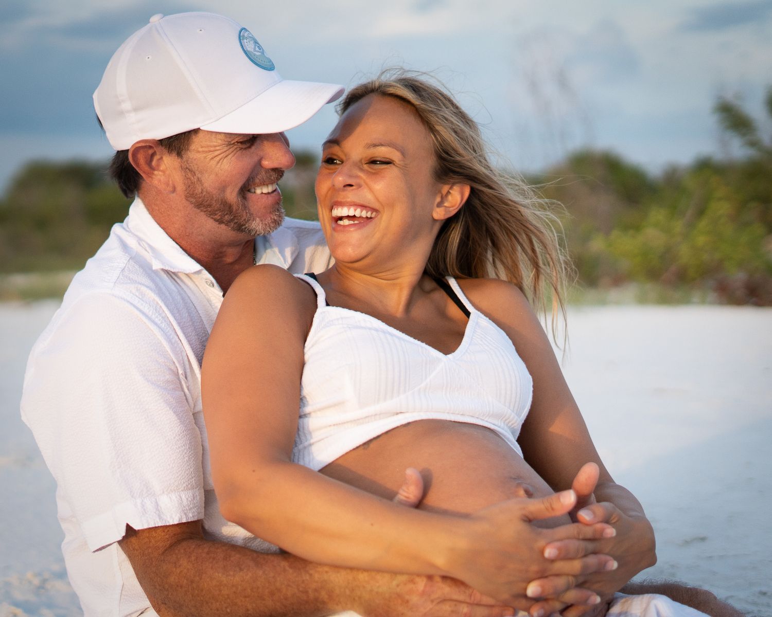 A romantic beach photoshoot scene with people in white clothing embracing against a bright sky.