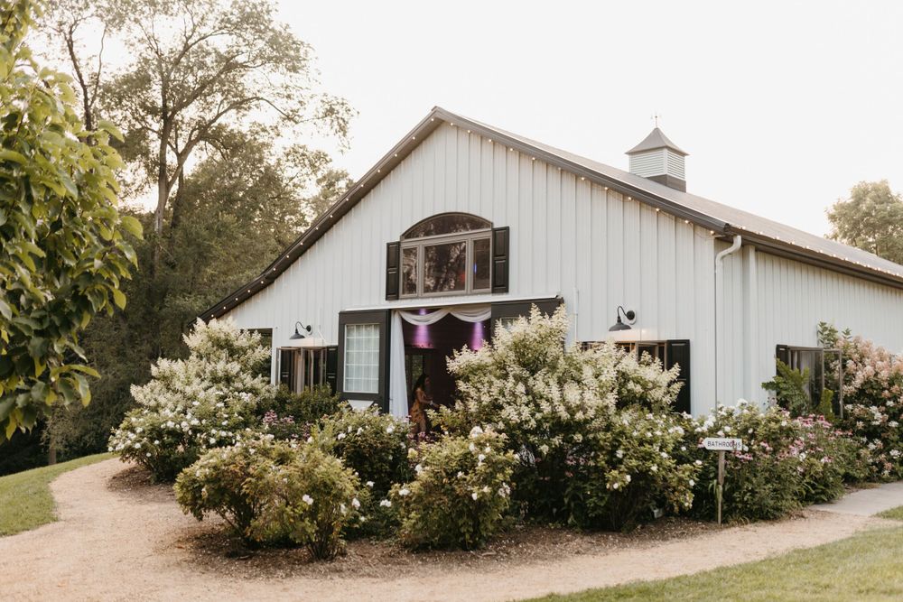 A charming white barn wedding venue surrounded by blooming bushes and lush trees.