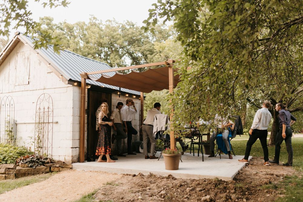 Wedding guests mingle on a rustic outdoor patio beside a white brick building.