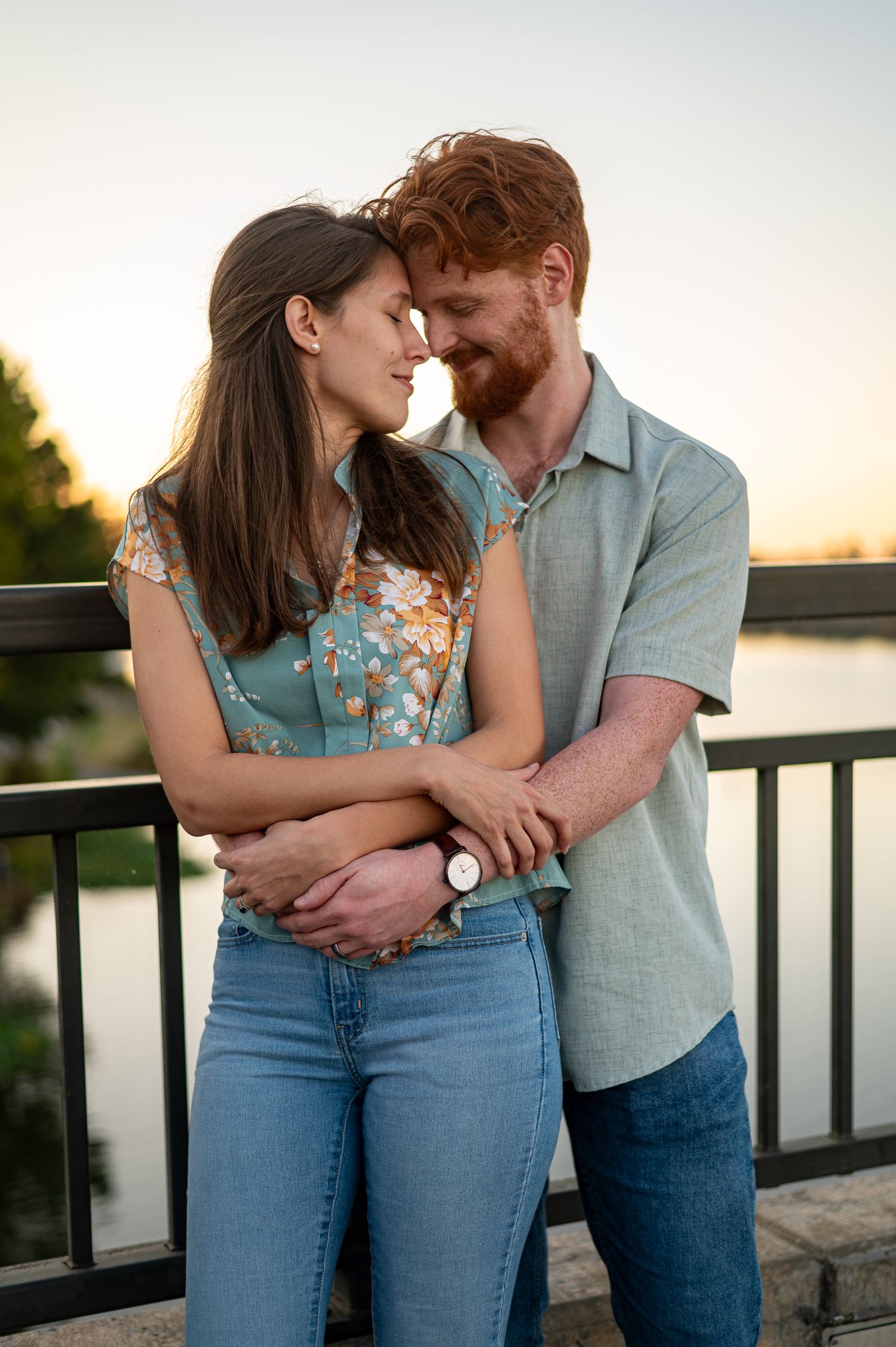 A couple with red and brown hair embrace tenderly on a bridge at sunset near a serene lake.