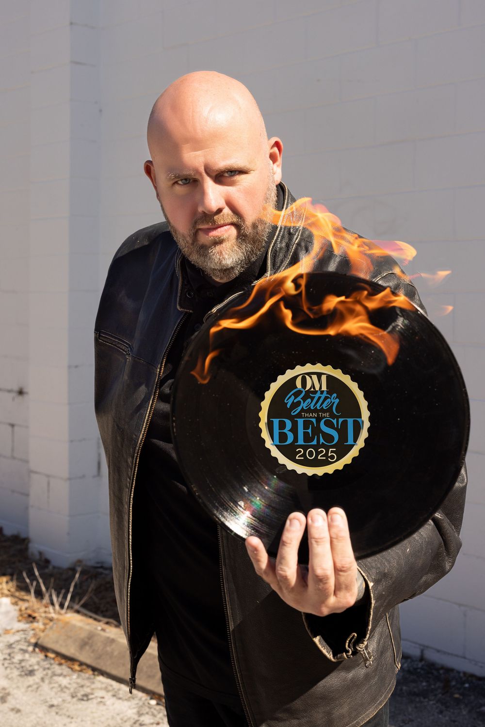 Bald man in leather jacket holding flaming vinyl record with OM Better Best 2025 award label.