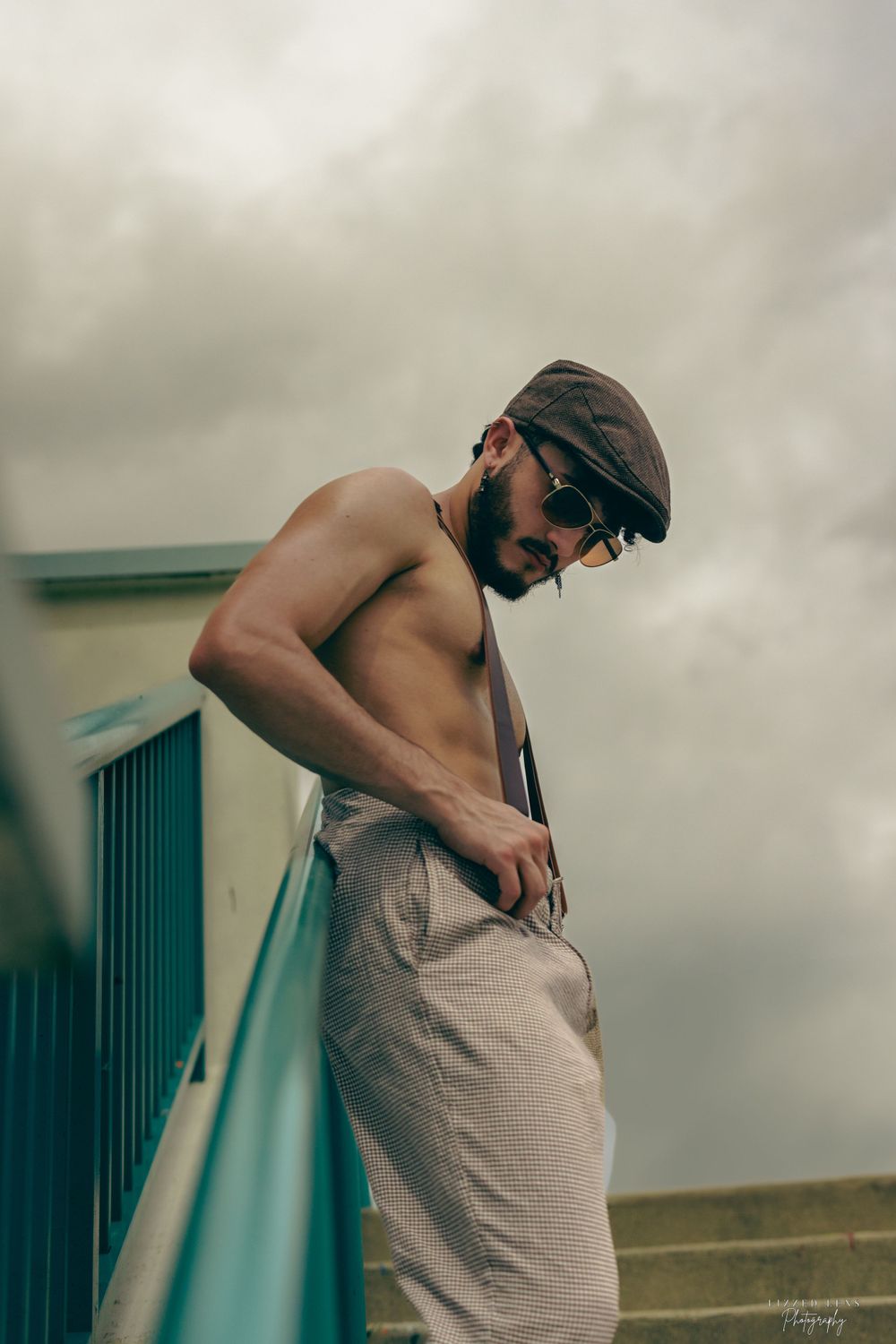 Shirtless bearded man in cap and sunglasses leaning on teal railing under cloudy sky.