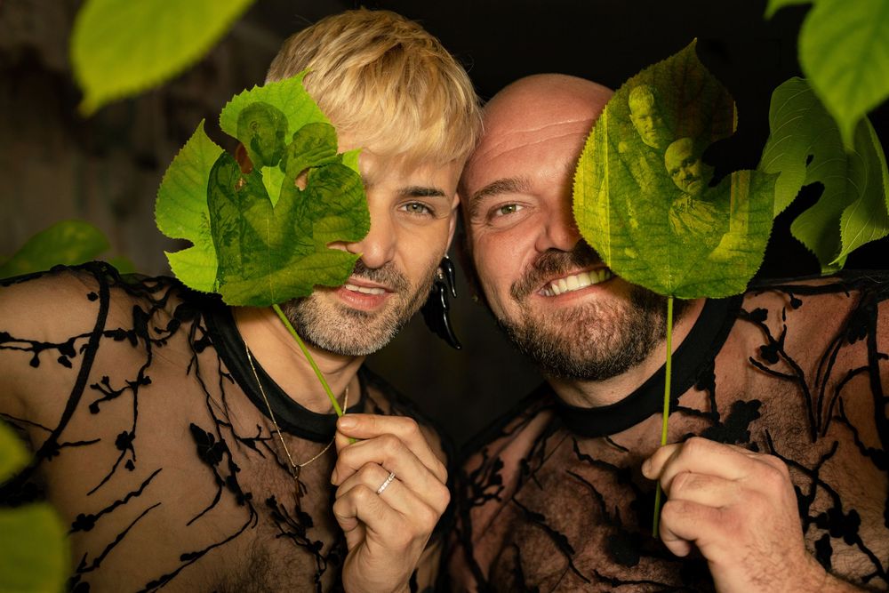 Two men playfully peek through large green leaves while wearing matching black floral sheer tops outdoors.