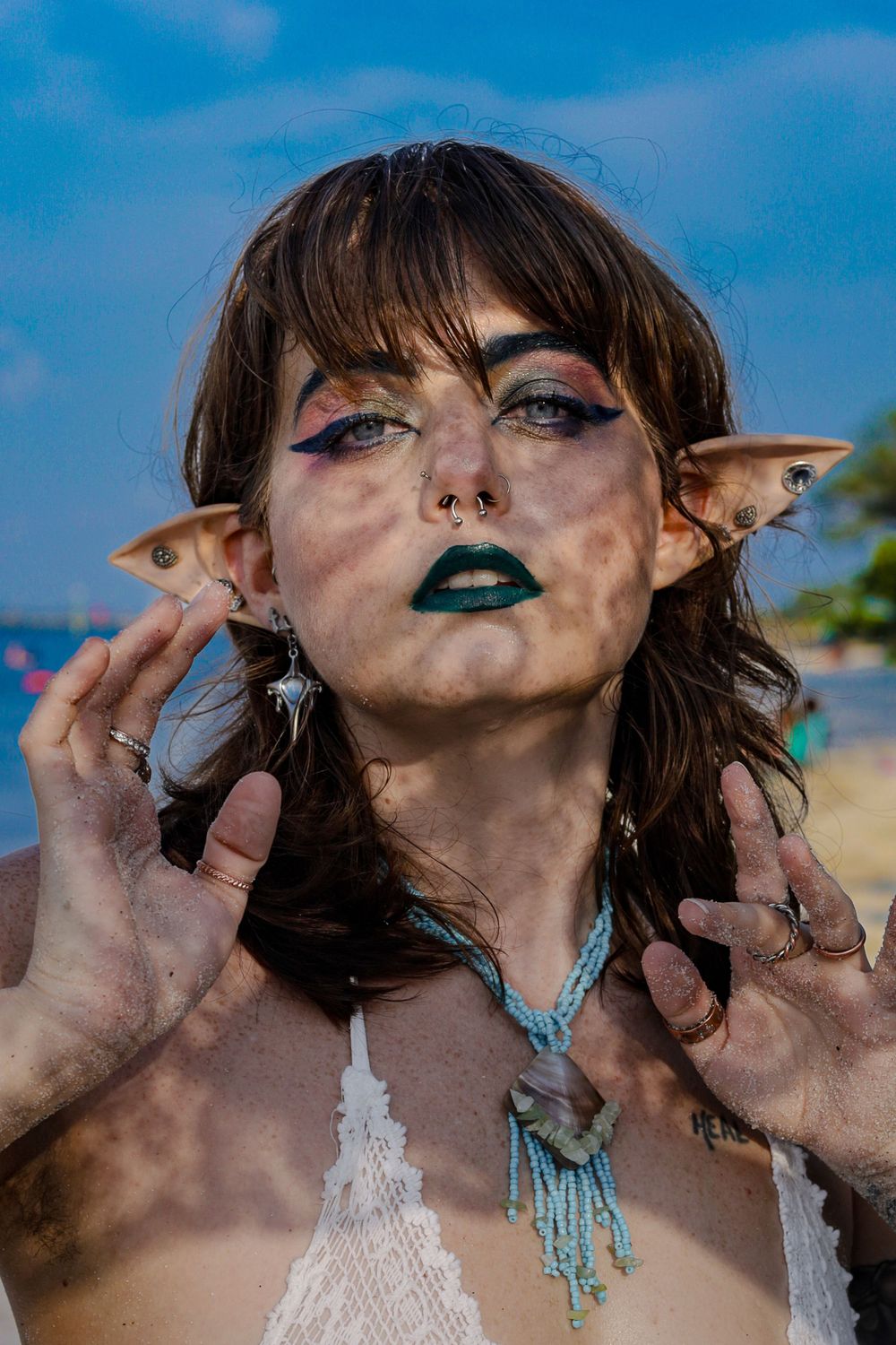 Woman with elf ears, green lipstick, vitiligo, and bohemian jewelry poses on a beach.