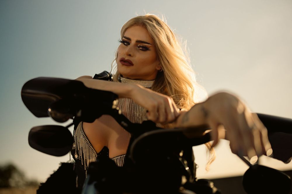 Blonde woman in black fringe jacket posing on motorcycle at sunset with dramatic sky background.