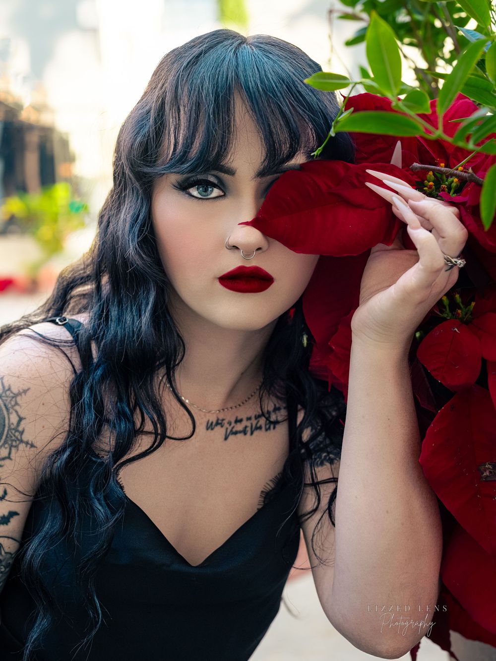 Dark-haired woman with bold red lips and tattoos poses beside vibrant red flowers in a dramatic portrait.
