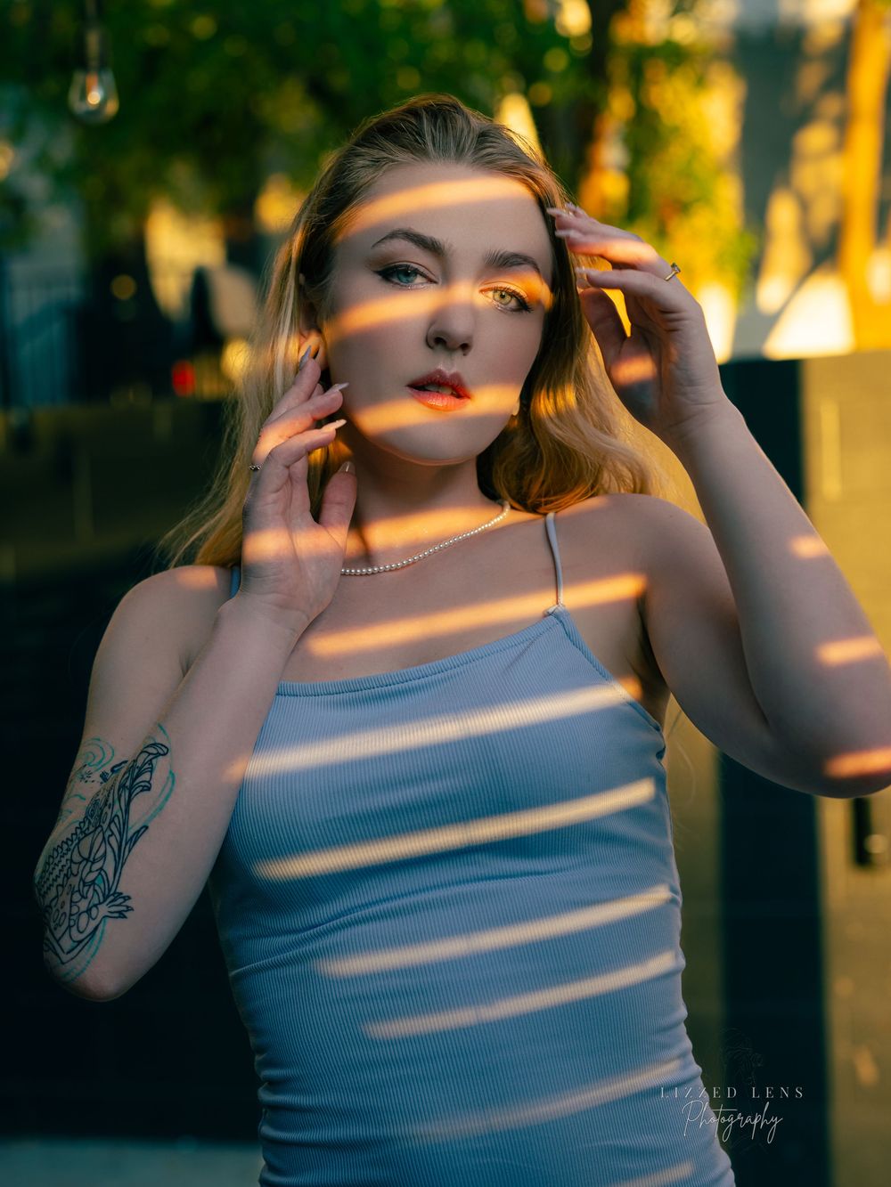 Young blonde woman with arm tattoo poses in golden hour light with artistic shadow stripes across her face.
