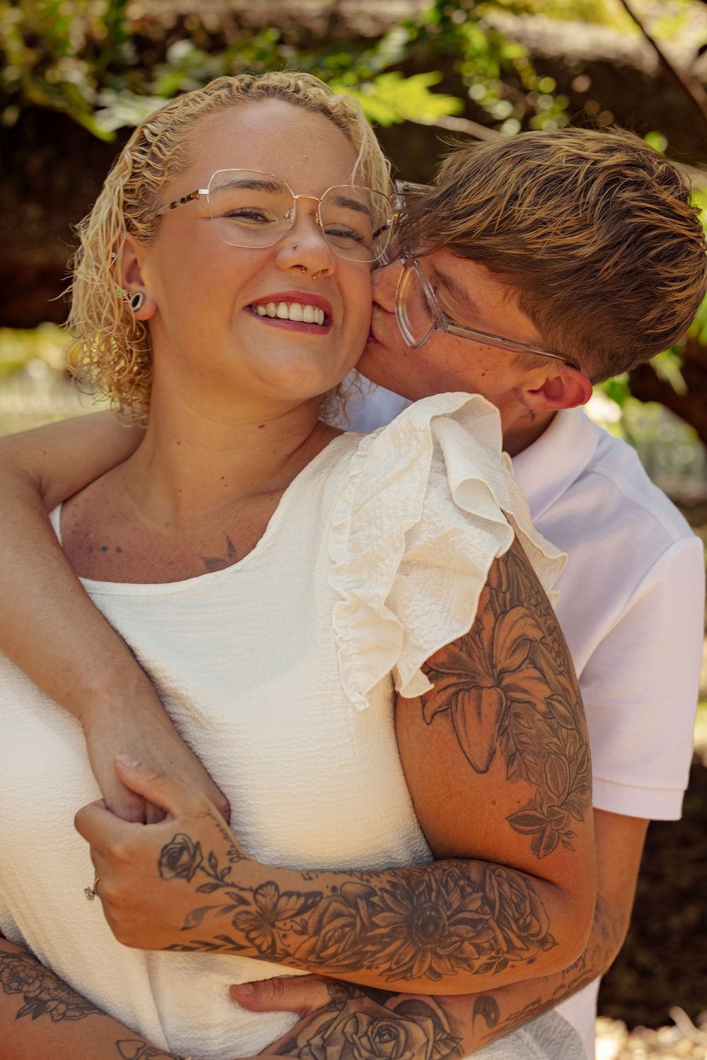 A couple embraces outdoors, woman in white dress smiling as tattooed partner kisses her cheek near green trees.