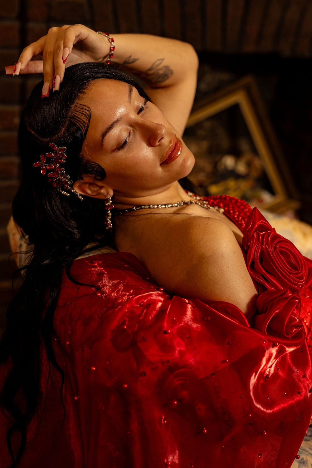 A woman in a red satin gown with rose details poses gracefully, wearing ruby jewelry and a hair accessory.