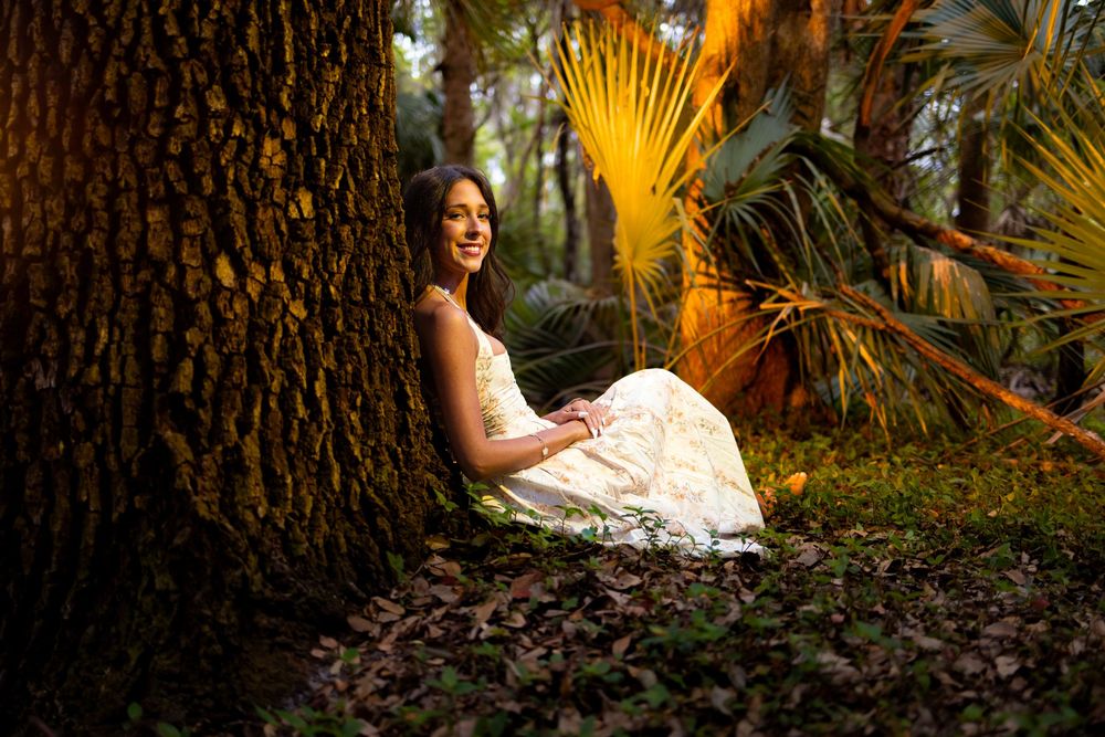 Young woman in white dress sitting peacefully against a large tree in a lush tropical forest.