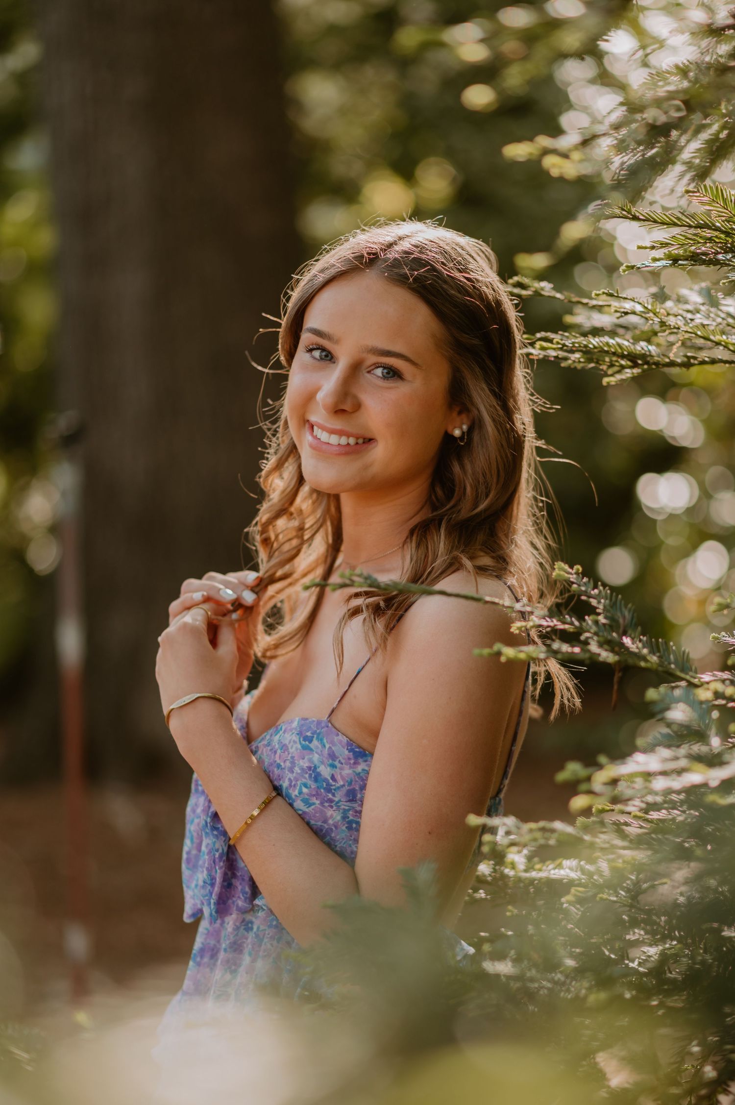 A senior girl in a floral dress smiles while standing among trees in a sunlit forest.