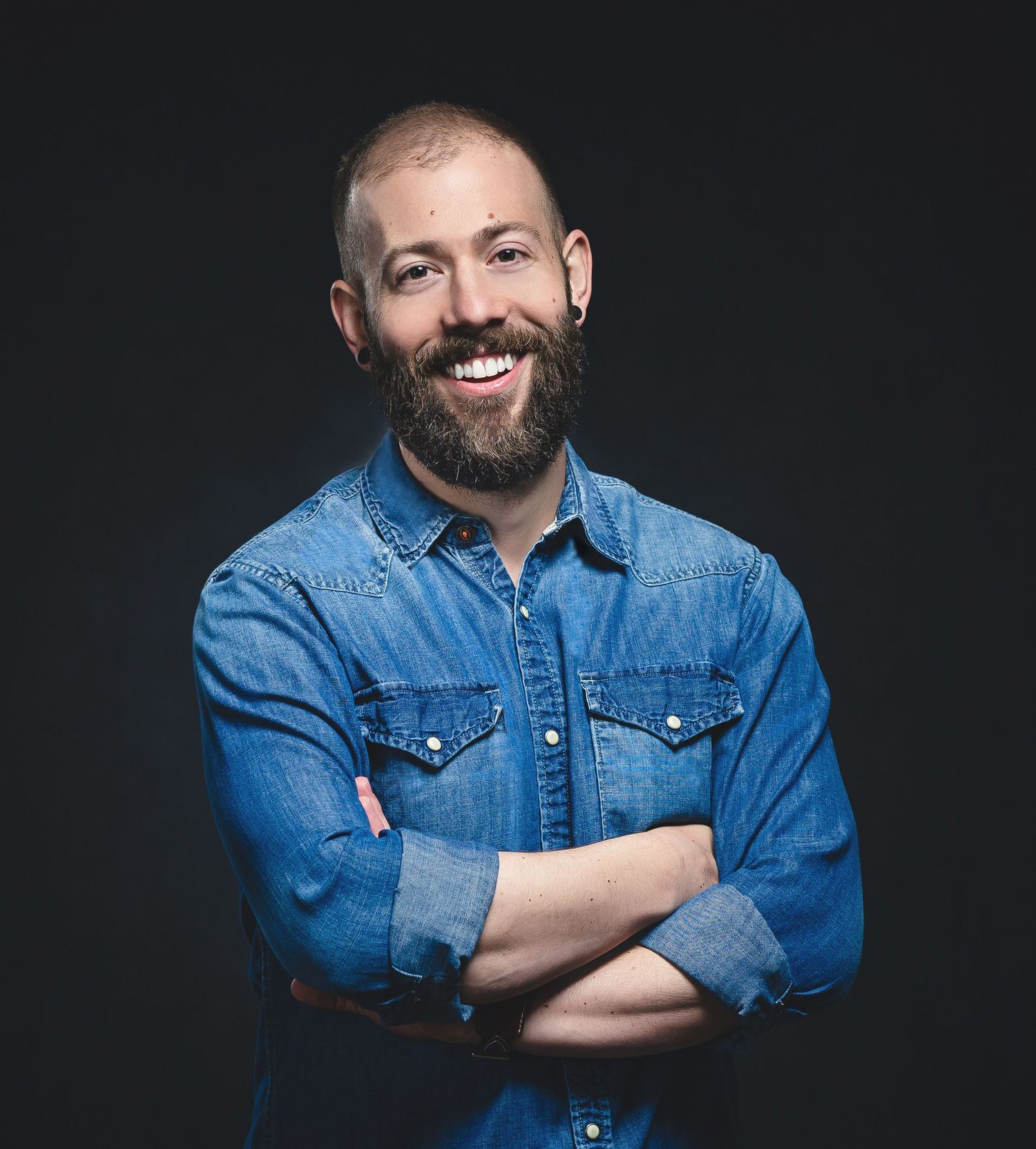 Portrait of person in denim shirt with beard against dark background.
