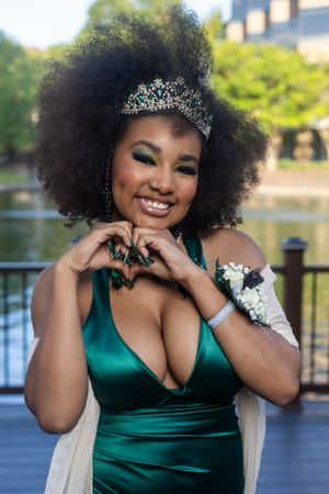 Formal portrait in emerald gown with tiara against outdoor waterfront setting.