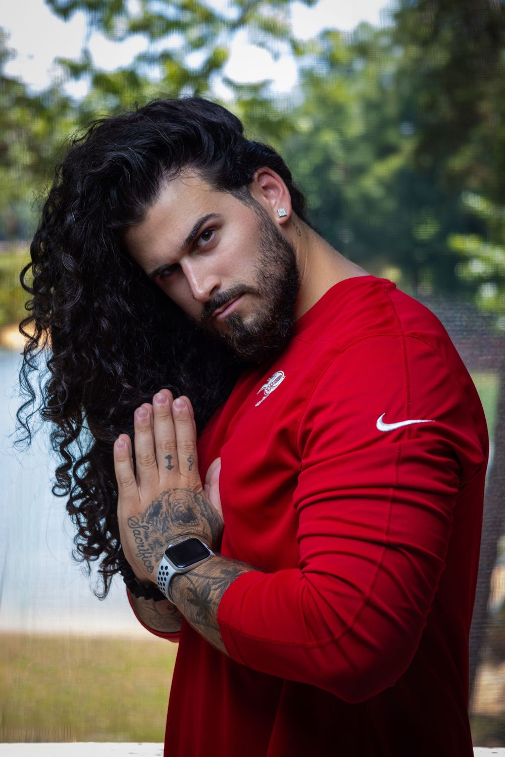 Athletic person in red Nike shirt poses outdoors with long dark curly hair and tattoos.
