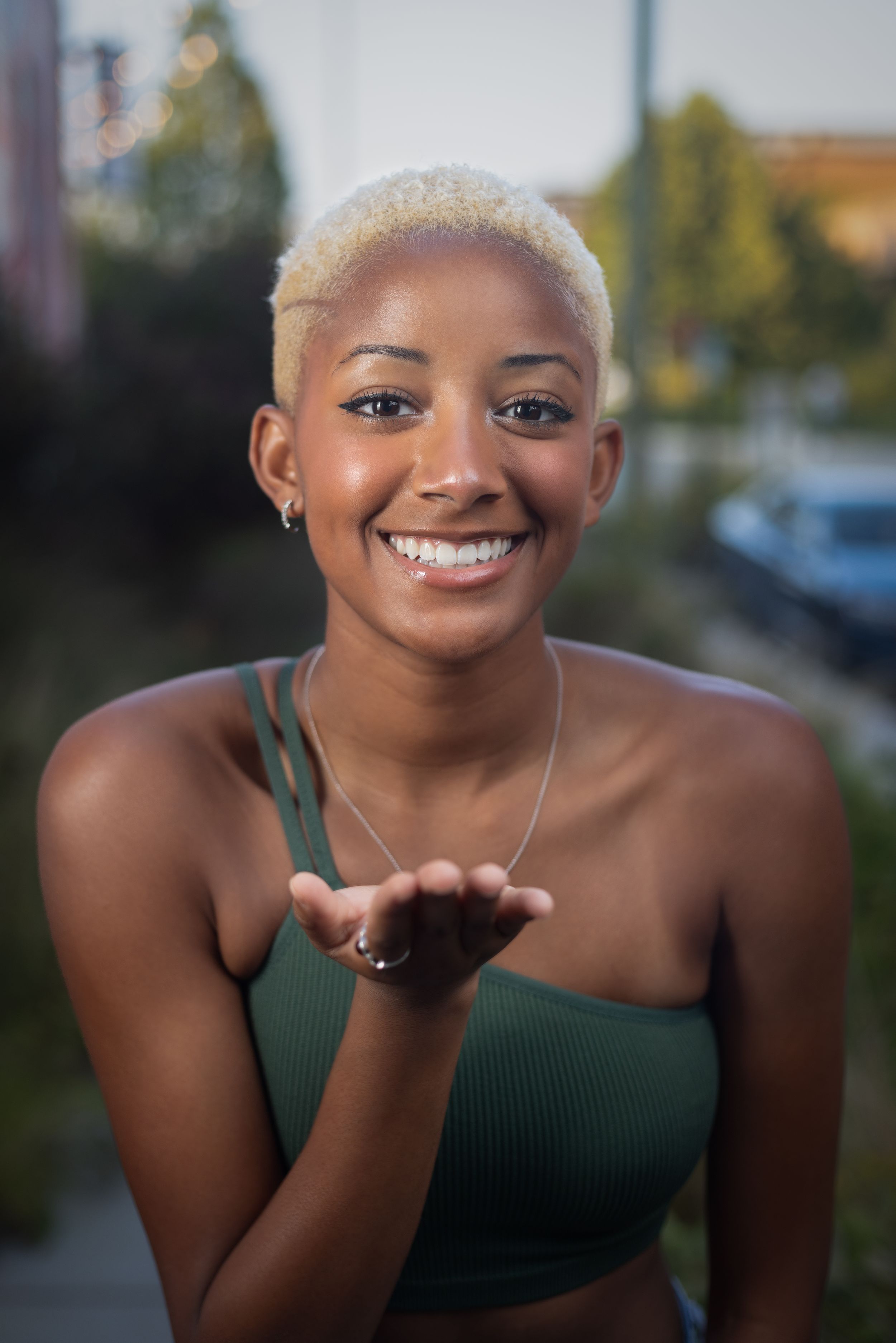 Portrait of someone with platinum blonde hair and bright smile blowing a kiss outdoors.