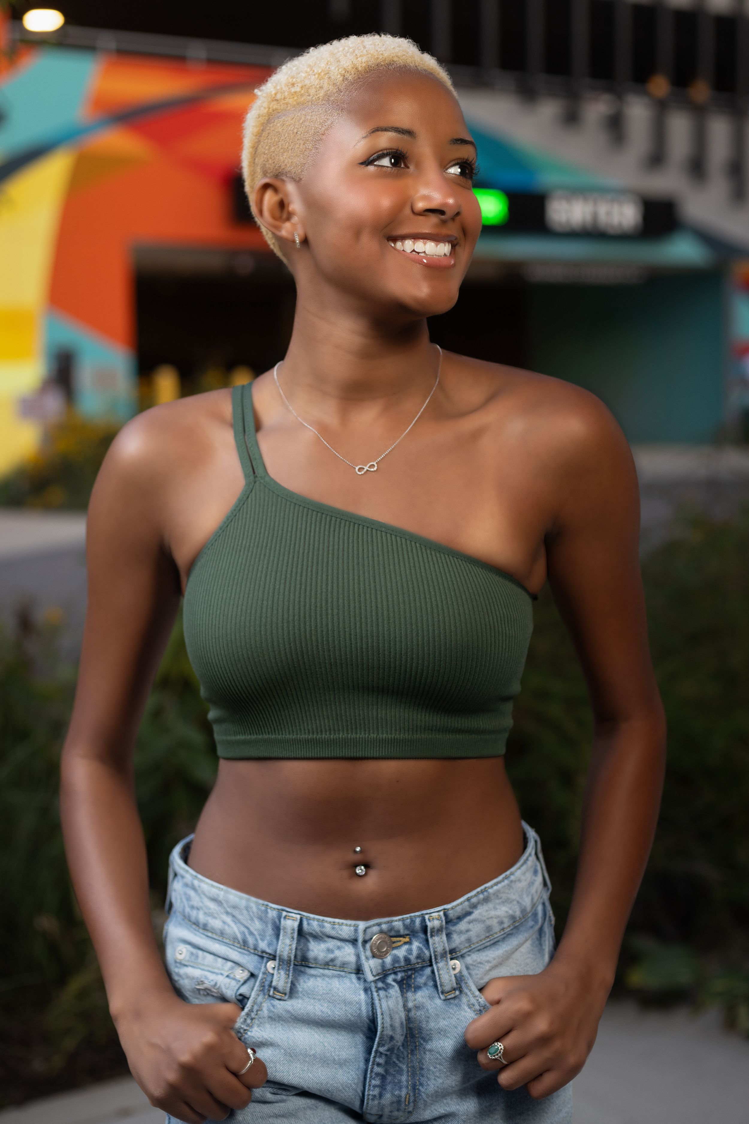 Portrait of someone in a green crop top and denim jeans posing outdoors near colorful wall art.