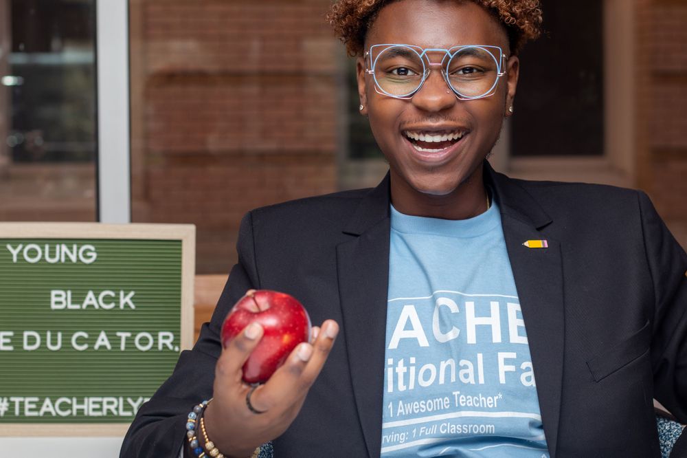 A smiling educator holds a red apple while wearing glasses and a blue t-shirt that says TEACHER.