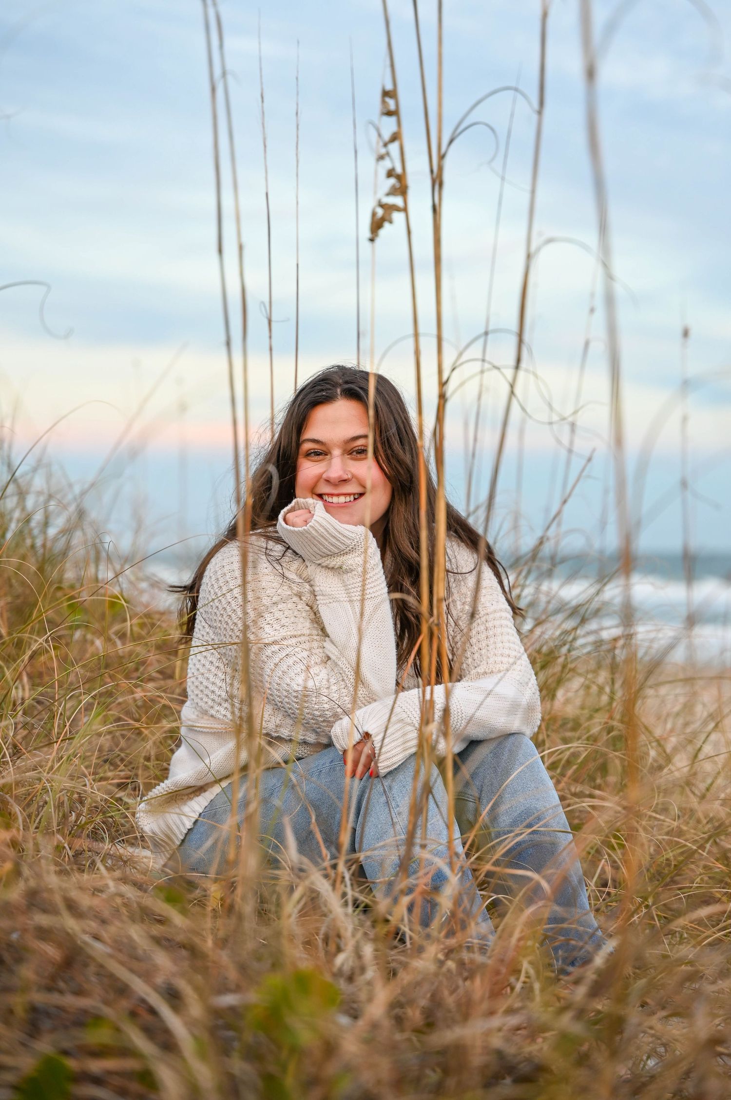 A person in a cozy sweater sits among beach grass during sunset.