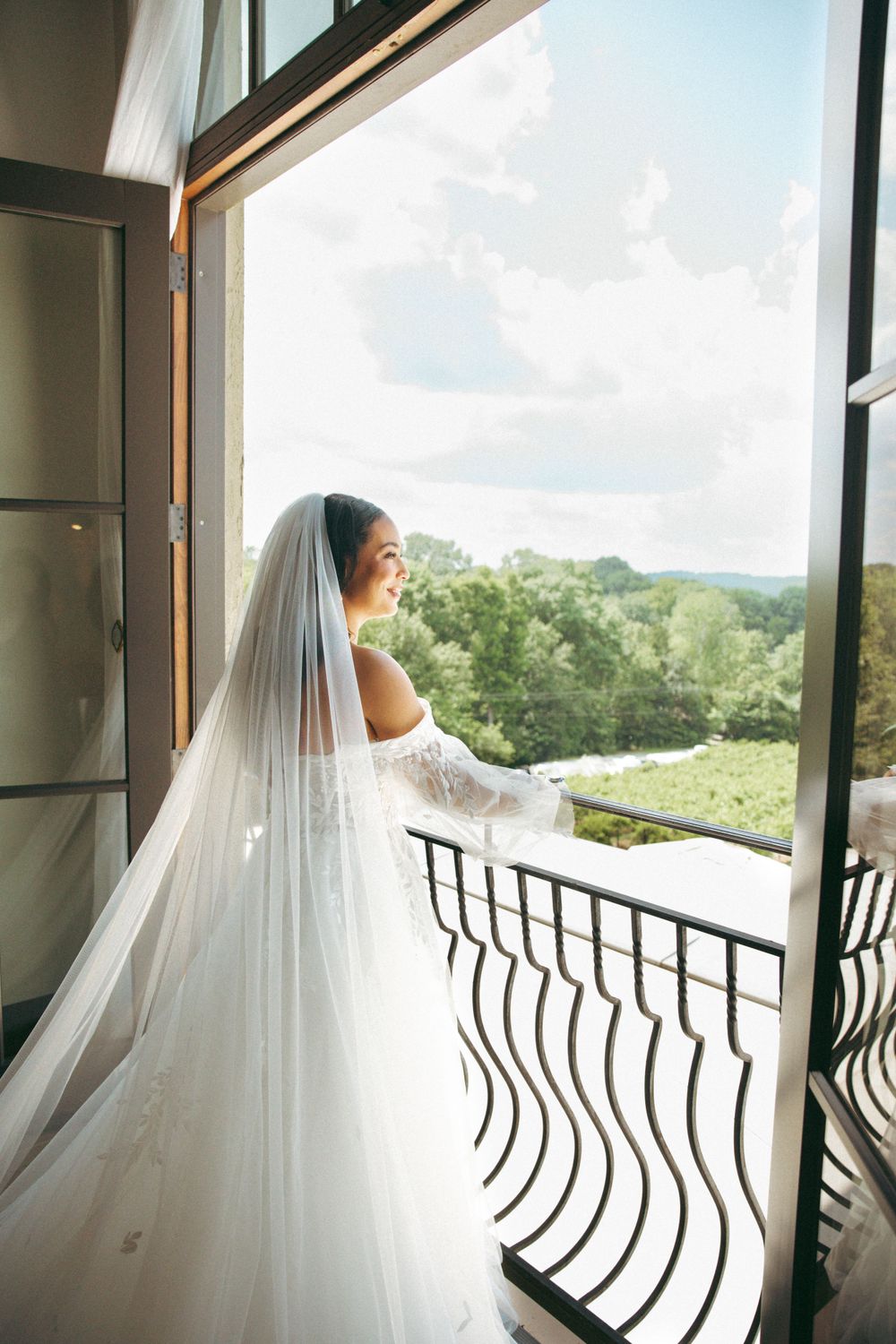 A bride in a flowing white wedding dress and veil stands on a balcony overlooking a scenic mountain landscape.