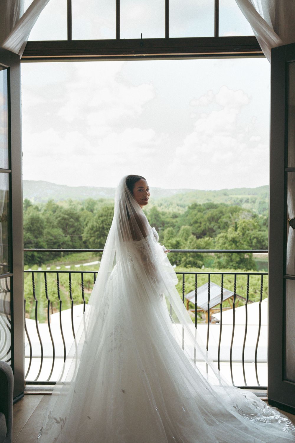 A bride silhouetted against large windows gazes at mountain views from a balcony on her wedding day.