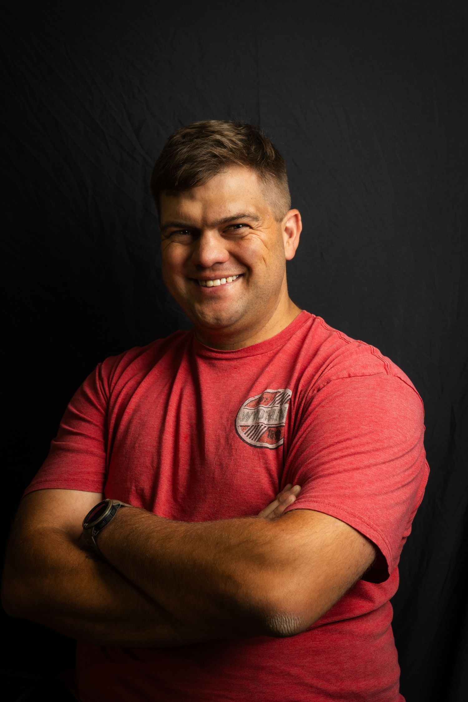 Professional studio portrait of a person wearing a red t-shirt with crossed arms and smiling against a dark background.
