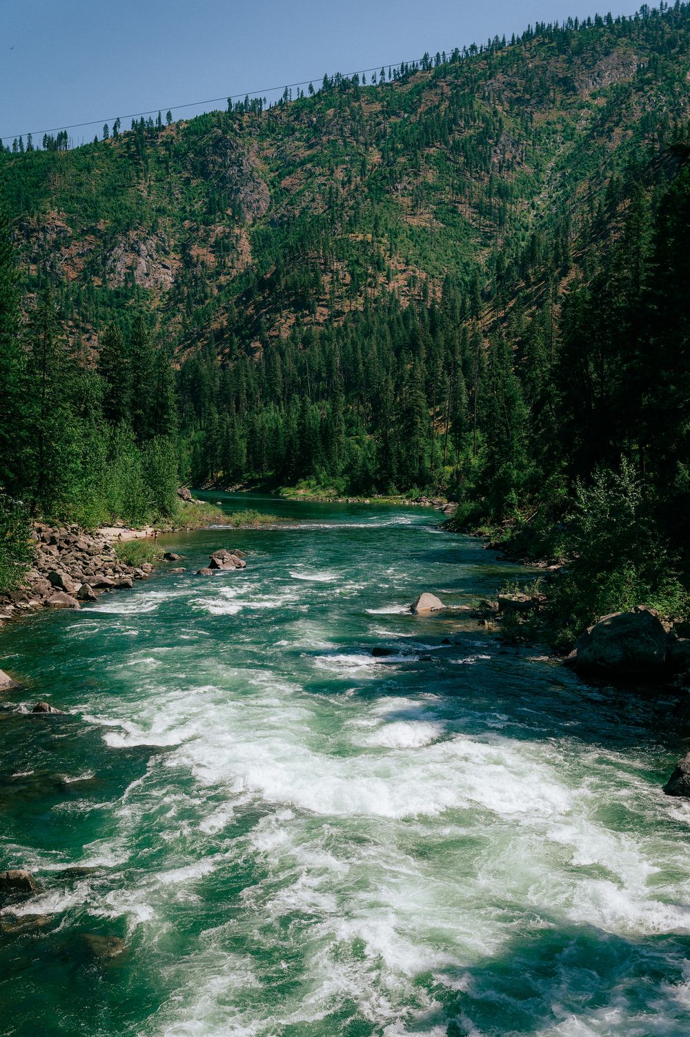 Rushing turquoise river waters flow through a mountainous forest landscape with steep rocky cliffs.