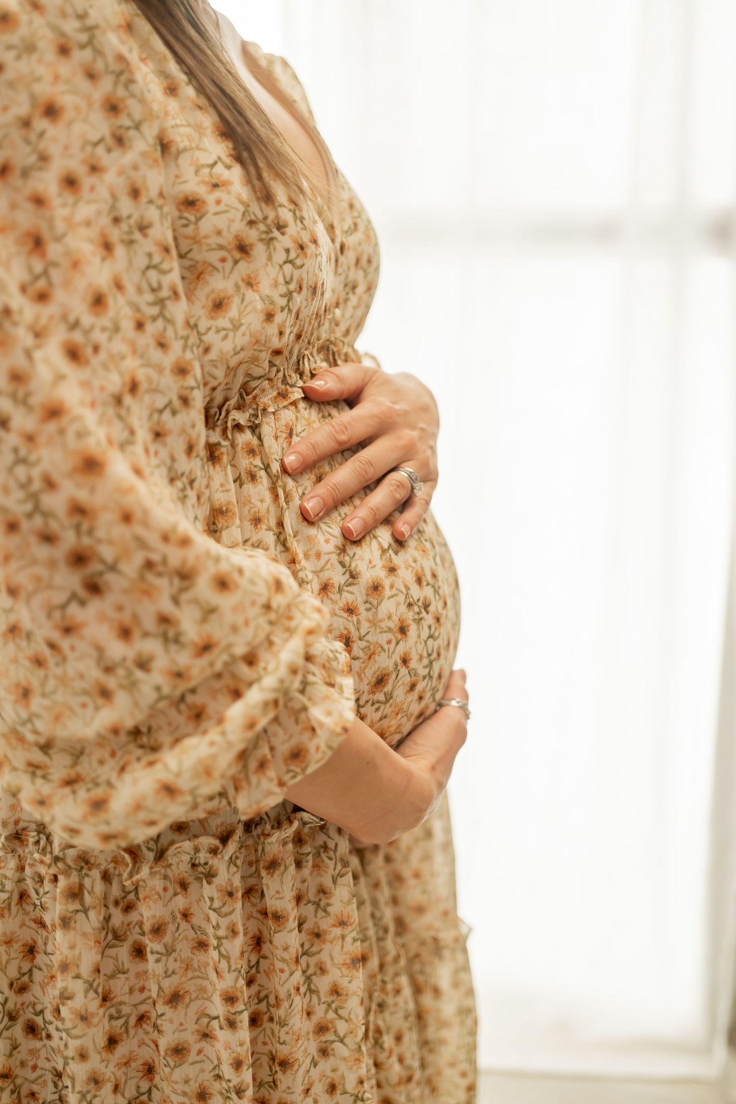 Close up of hands cradling pregnant belly in floral dress against soft white background.