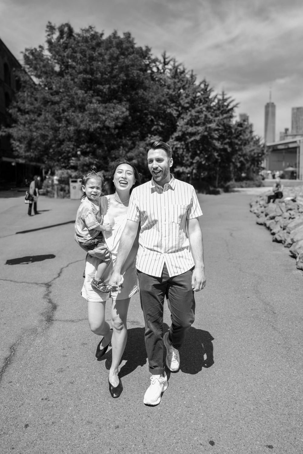 A family walks together on a sunny day in a city park with urban buildings visible in the background.