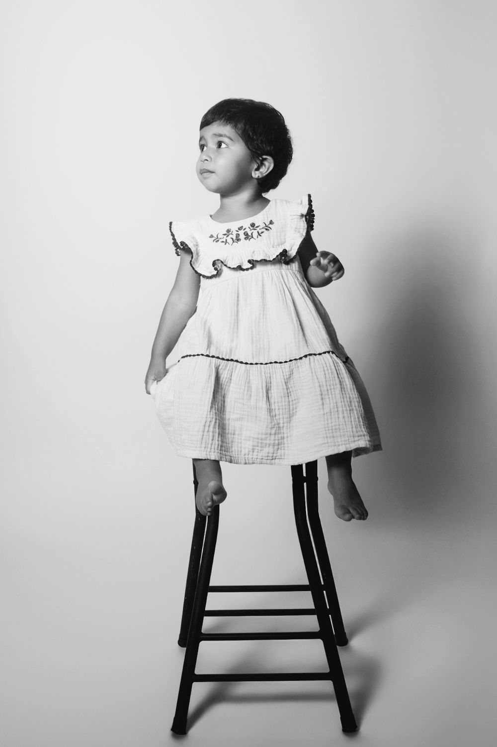 Black and white artistic composition of a small child wearing a white dress perched on a dark wooden stool.