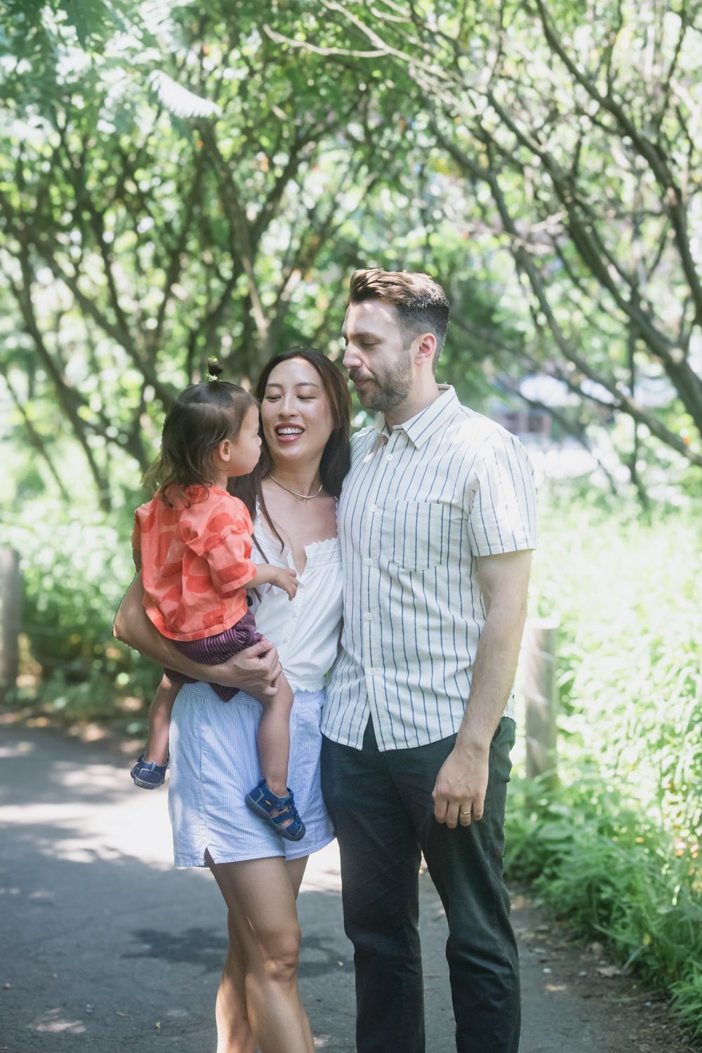 A couple and young child walk along a tree-lined path on a sunny summer day, shown in four candid moments together.