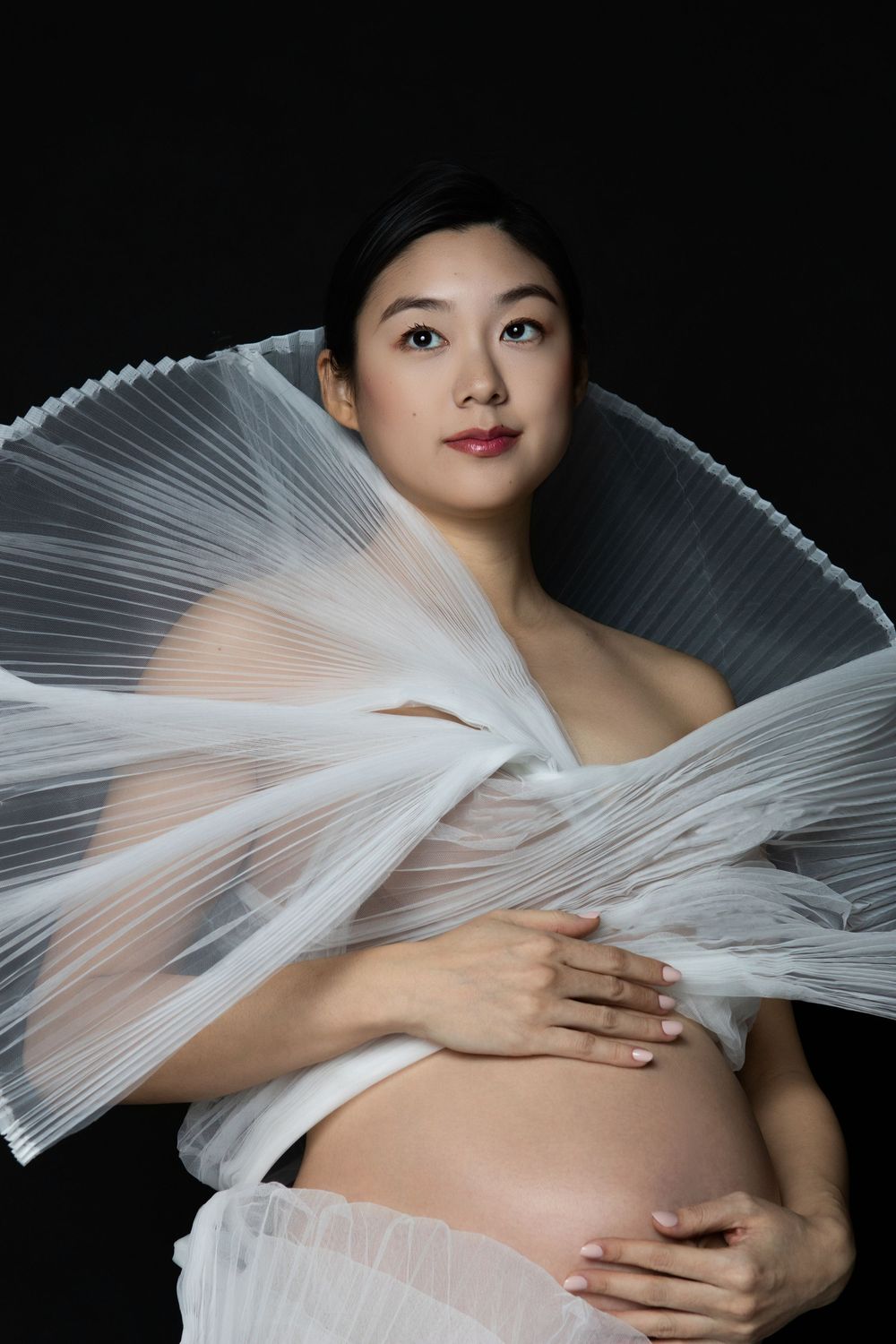 Pregnant Asian woman cradling her belly, draped in white pleated fabric against a black background.