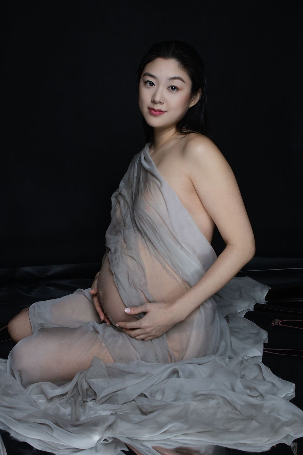 Pregnant woman in sheer gray fabric poses gracefully against dark background, cradling her baby bump in a maternity photo.