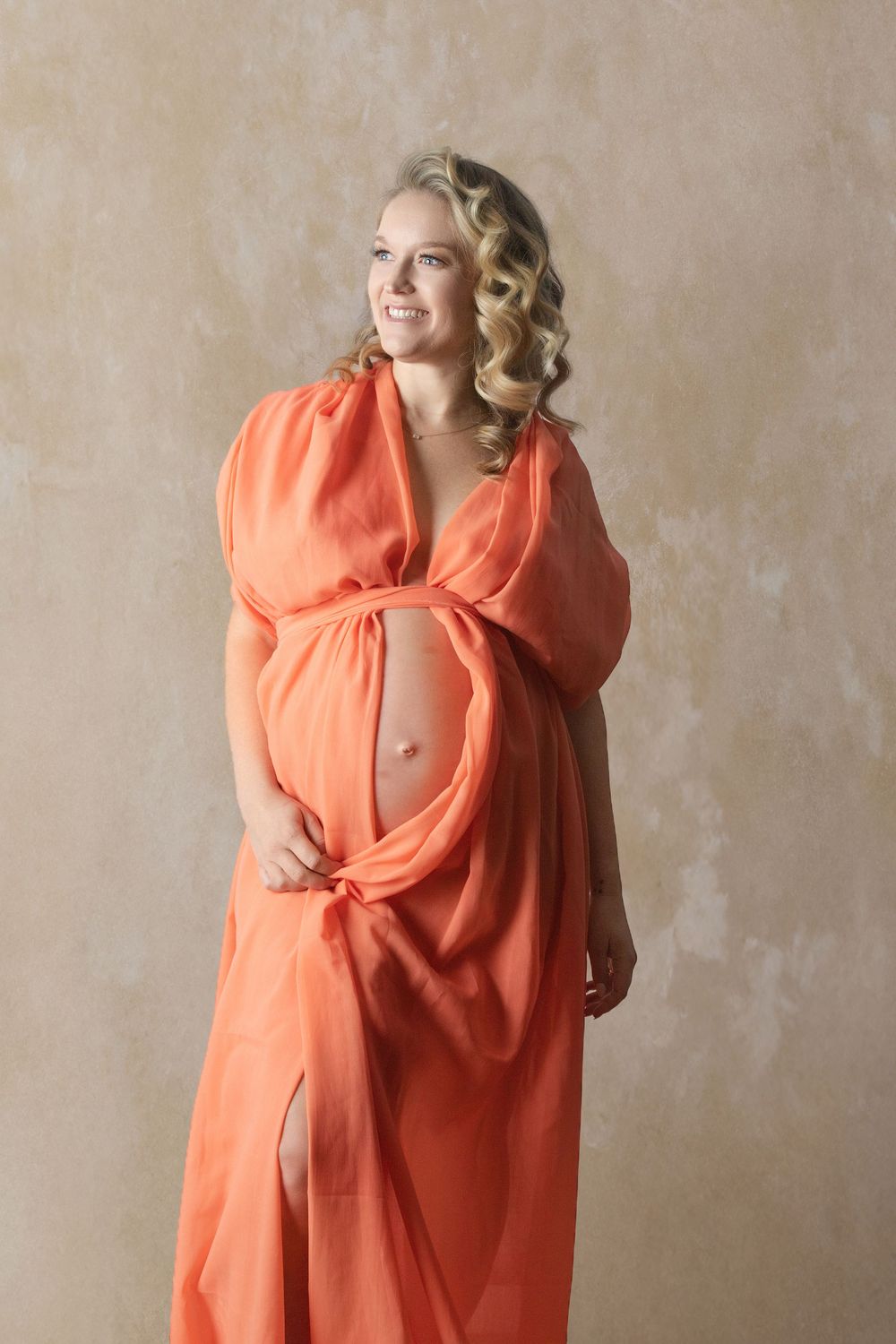 Smiling pregnant woman in flowing orange fabric dress posing gracefully, showing baby bump, with curly blonde hair.