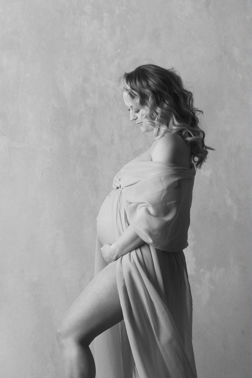 Black and white maternity photo of a pregnant woman in a flowing sheer gown, posing gracefully against a textured wall.